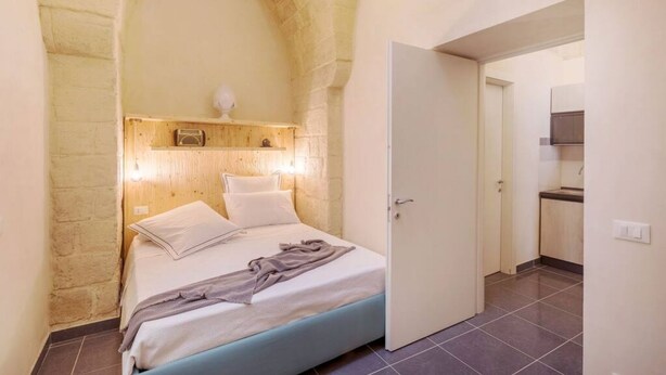 A bedroom with a stone archway, a bed with white linens, a bedside table with a lamp, and a view into a kitchen area. - Balbo 9A Apartment - Lecce Selection (Lecce)