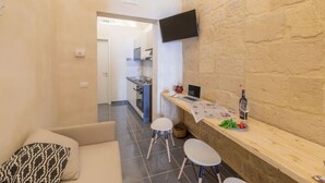 A compact living space with a kitchenette, a flat-screen TV mounted on a brick wall, and a small bar area with stools and a bottle of wine. - Balbo 9A Apartment - Lecce Selection (Lecce)