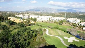 Eine Vogelperspektive auf einen Golfplatz, im Hintergrund ein großer, weißer Gebäudekomplex, umgeben von viel Grün und Bergen.