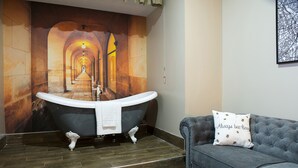 A bathroom with a freestanding bathtub, a grey sofa with a decorative pillow, and a tiled floor. - Manchester Hall (Manchester)