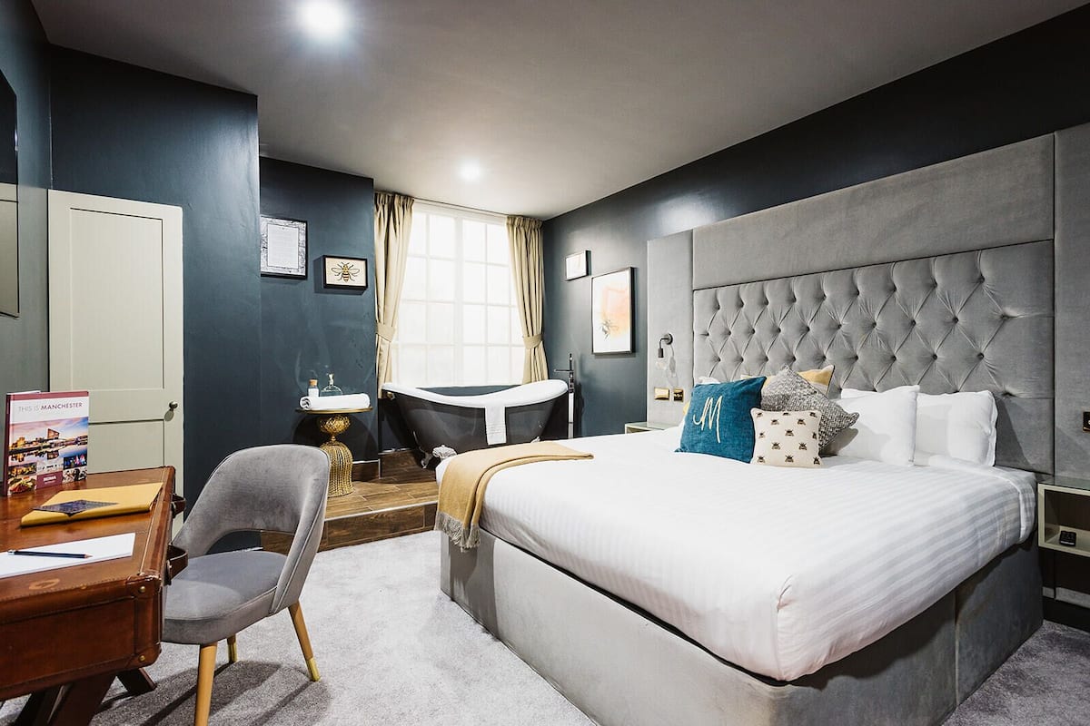 A modern hotel room with a large bed, a desk, a chair, and a vanity with a mirror.