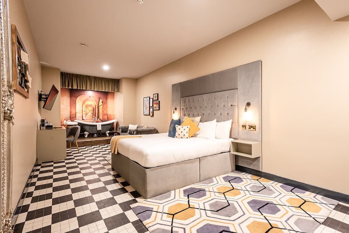 A hotel room with a checkered floor, a bed with multiple pillows, a desk with a chair, and a wall-mounted TV.