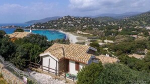 A coastal view with a building featuring a tiled roof, surrounded by greenery and a clear blue sea.