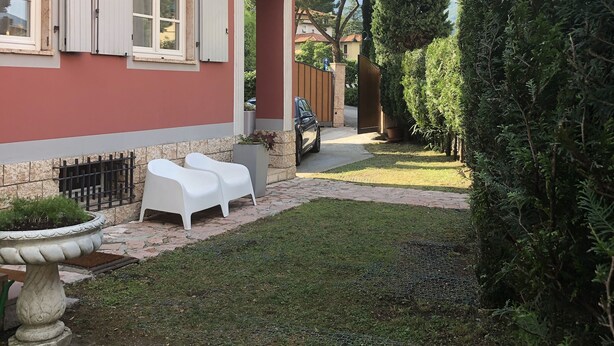 A red building with white shutters, a paved patio with two white chairs, and a car parked in the background.