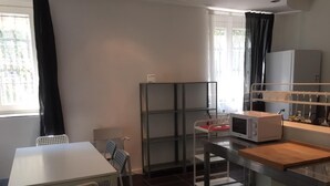 A compact kitchen with a microwave, a small table, and chairs. There is a window with curtains, a shelf unit, and a refrigerator.