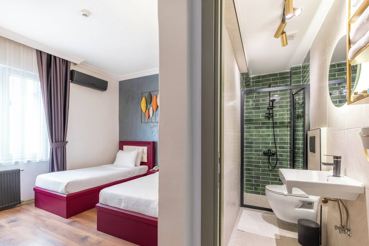 A hotel room with a bed, a bedside table, a window with curtains, a shower area with a glass door, and a bathroom with a sink and toilet.