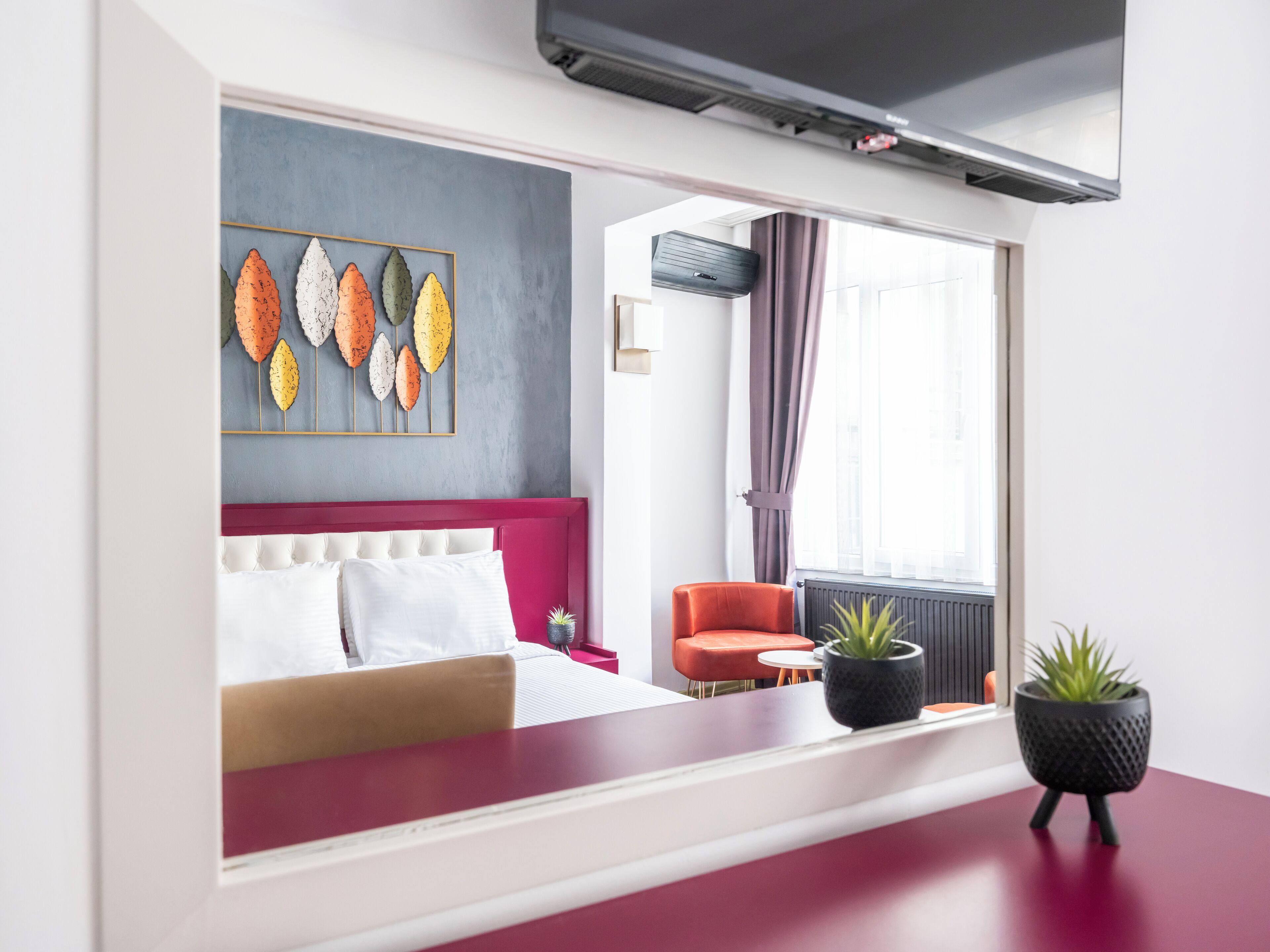 Ein modernes Hotelzimmer mit einem roten Sofa, einem weißen Bett, einer farbenfrohen Blattform-Kunst, einem roten Sessel und einer Topfpflanze.