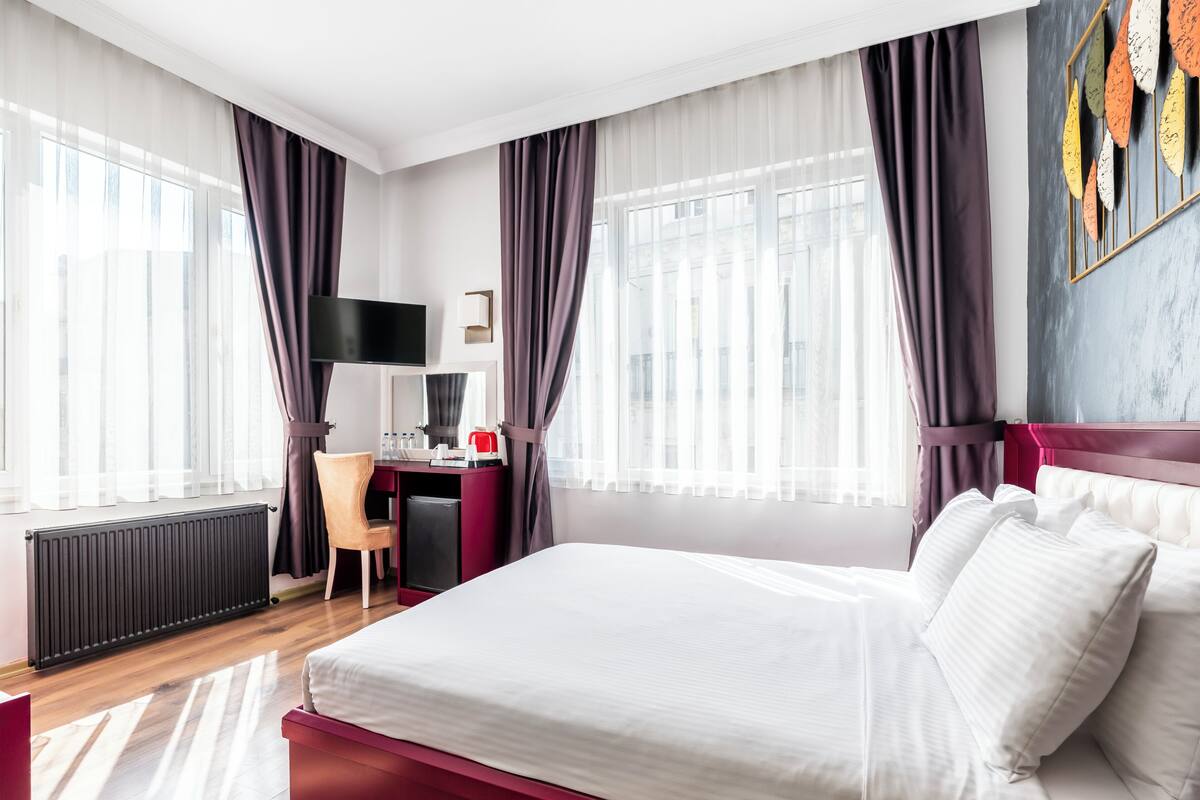 A hotel room with a bed, a desk, a chair, a TV, and a radiator.