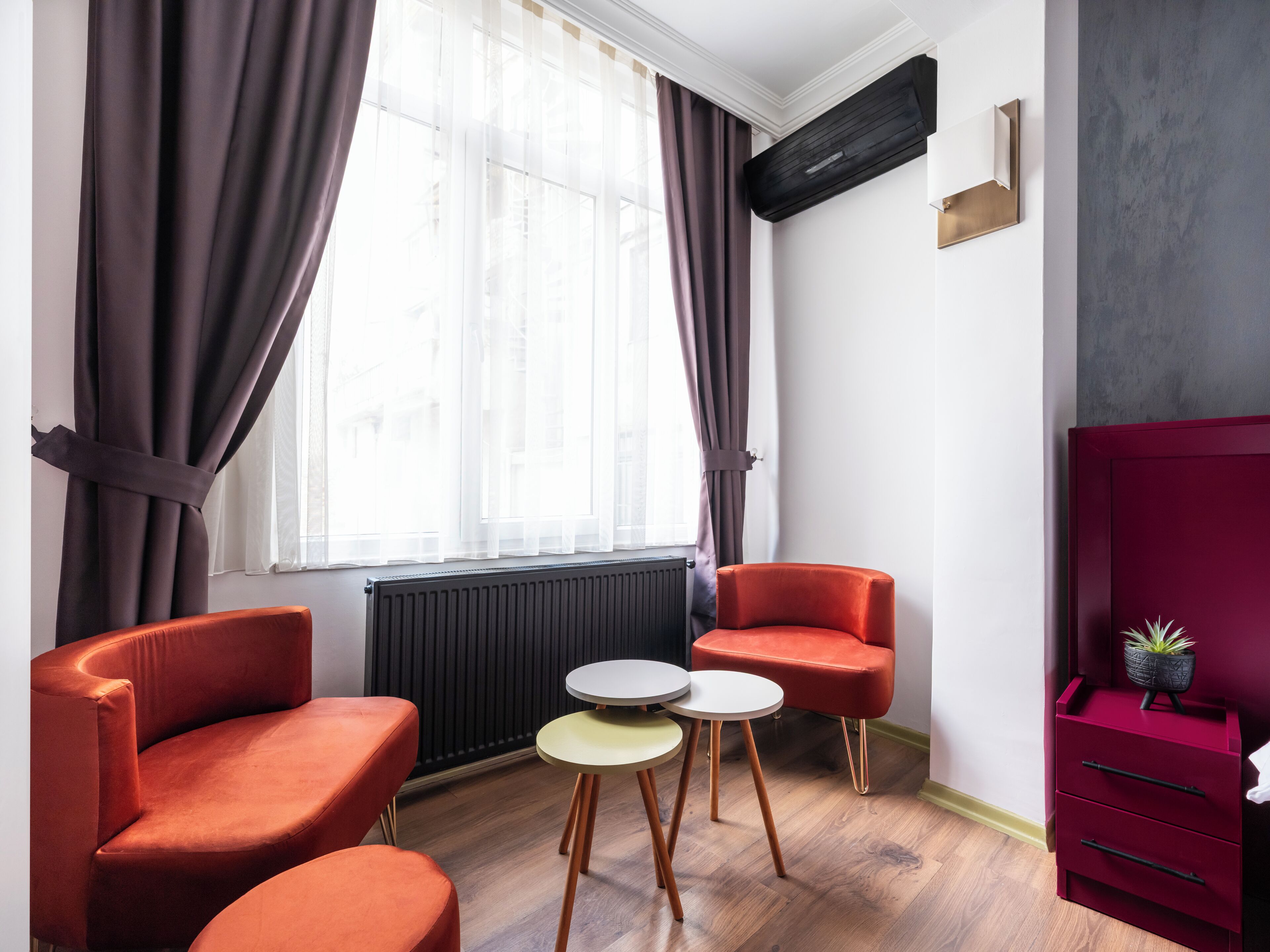 Habitació moderna amb dues cadires taronges, una taula rodona petita, un radiador, una finestra amb cortines i un armari vermell.