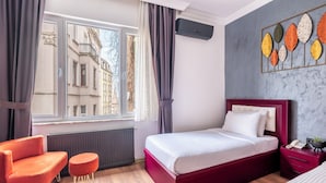 Une chambre d’hôtel avec un lit, une chaise, une petite table, un radiateur, un appareil de climatisation et une fenêtre avec des rideaux.