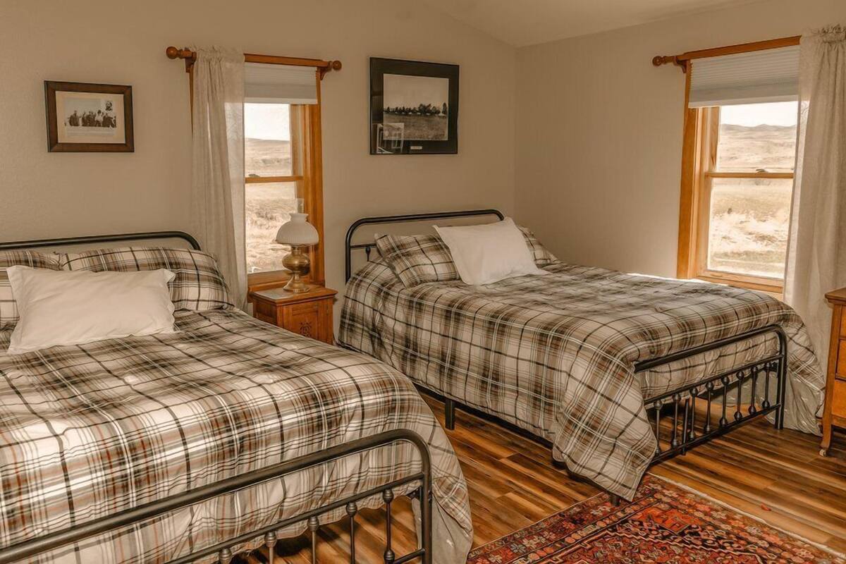 A room with two beds, each with a metal frame and plaid bedding. There are two bedside tables with lamps, a framed picture on the wall, and a window with curtains.