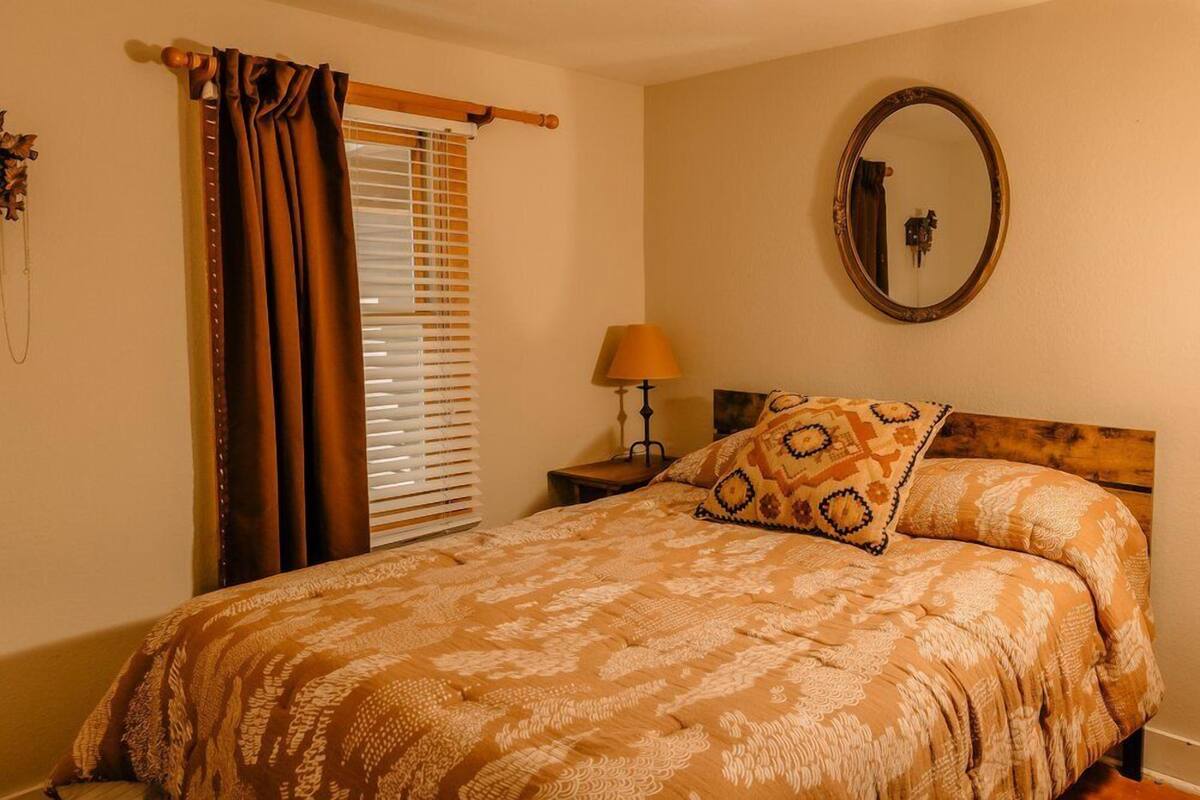 A bedroom with a bed, a nightstand, a lamp, a window with blinds, and a mirror on the wall.