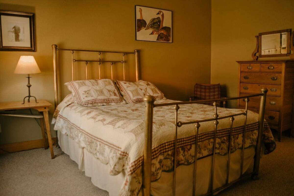 A bedroom with a metal bed frame, a wooden nightstand with a lamp, a wooden dresser, and a framed picture on the wall.