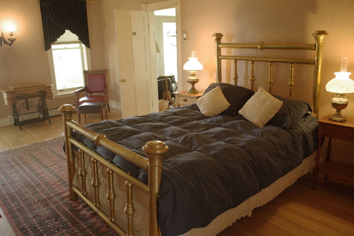 A neatly made bed with a dark blue comforter and two pillows, a brass headboard, a small bedside table with a lamp, and a patterned rug on the floor.