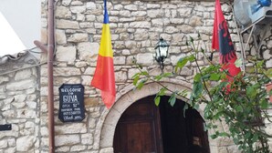 A stone building with a wooden door, flags, and a welcome sign. - Guva Mangalem (Berat)
