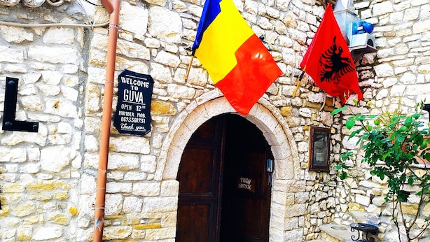 A stone building with a wooden door, two flags, and a welcome sign. - Guva Mangalem (Berat)