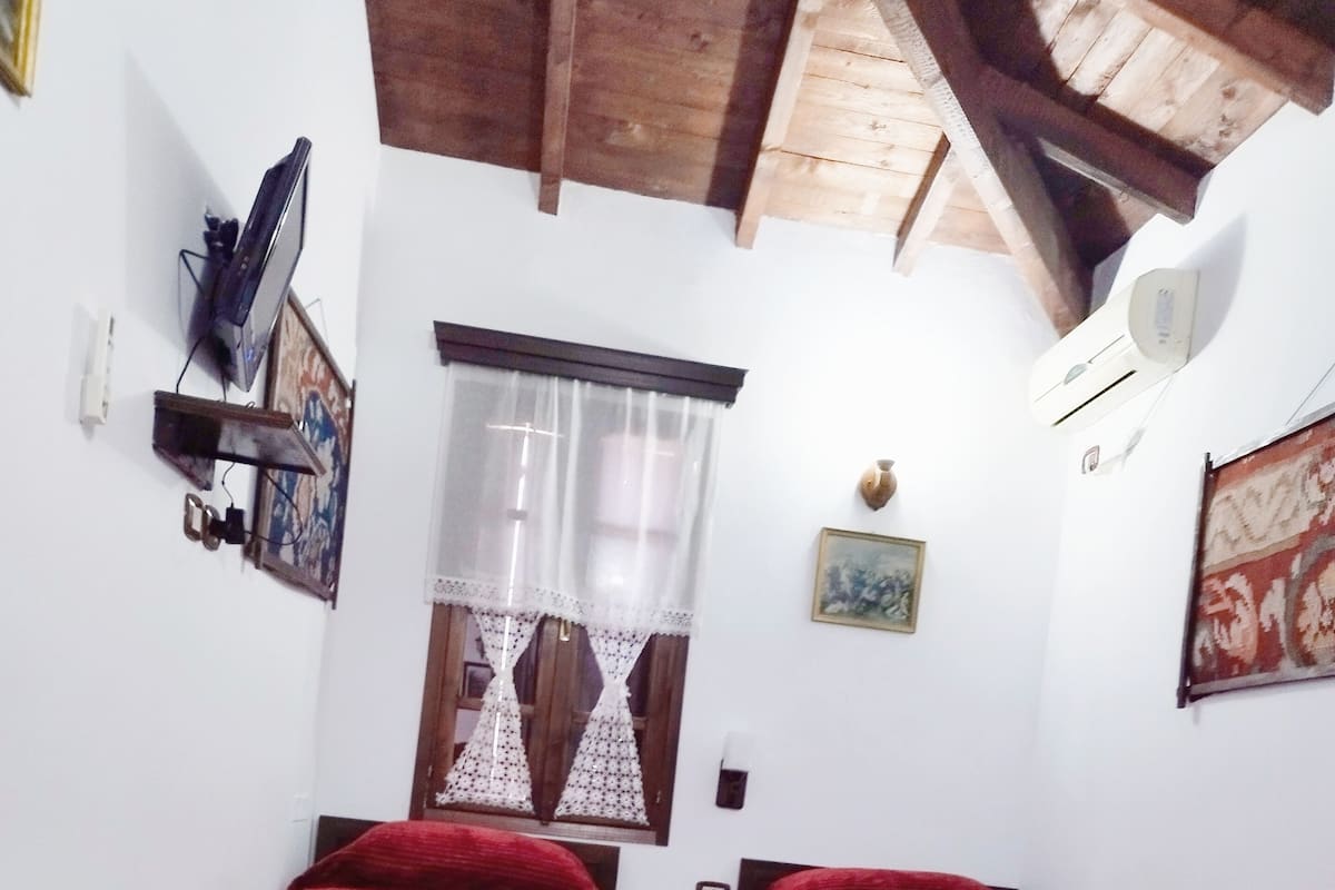 A small, cozy room with two single beds, a wooden table, a chair, a window with lace curtains, and a ceiling fan.