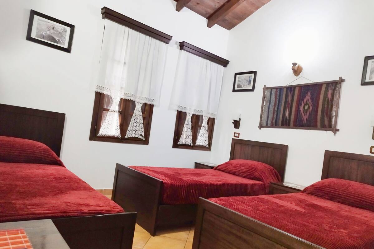 A room with three wooden beds, each with a red blanket, white curtains with lace details, and a wooden headboard. There are framed pictures on the walls, a colorful tapestry, and a small table with a checkered tablecloth.