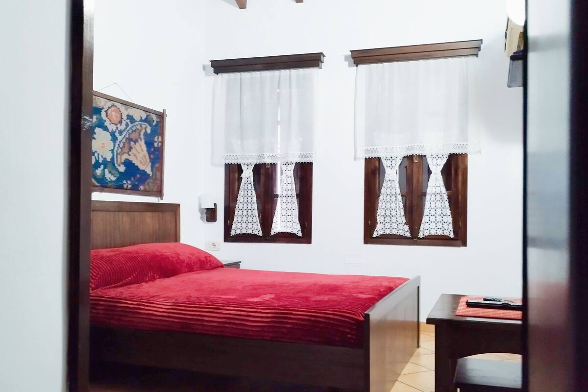 A bedroom with a wooden ceiling, two windows with lace curtains, a bed with a red blanket, a small table, and a patterned rug.