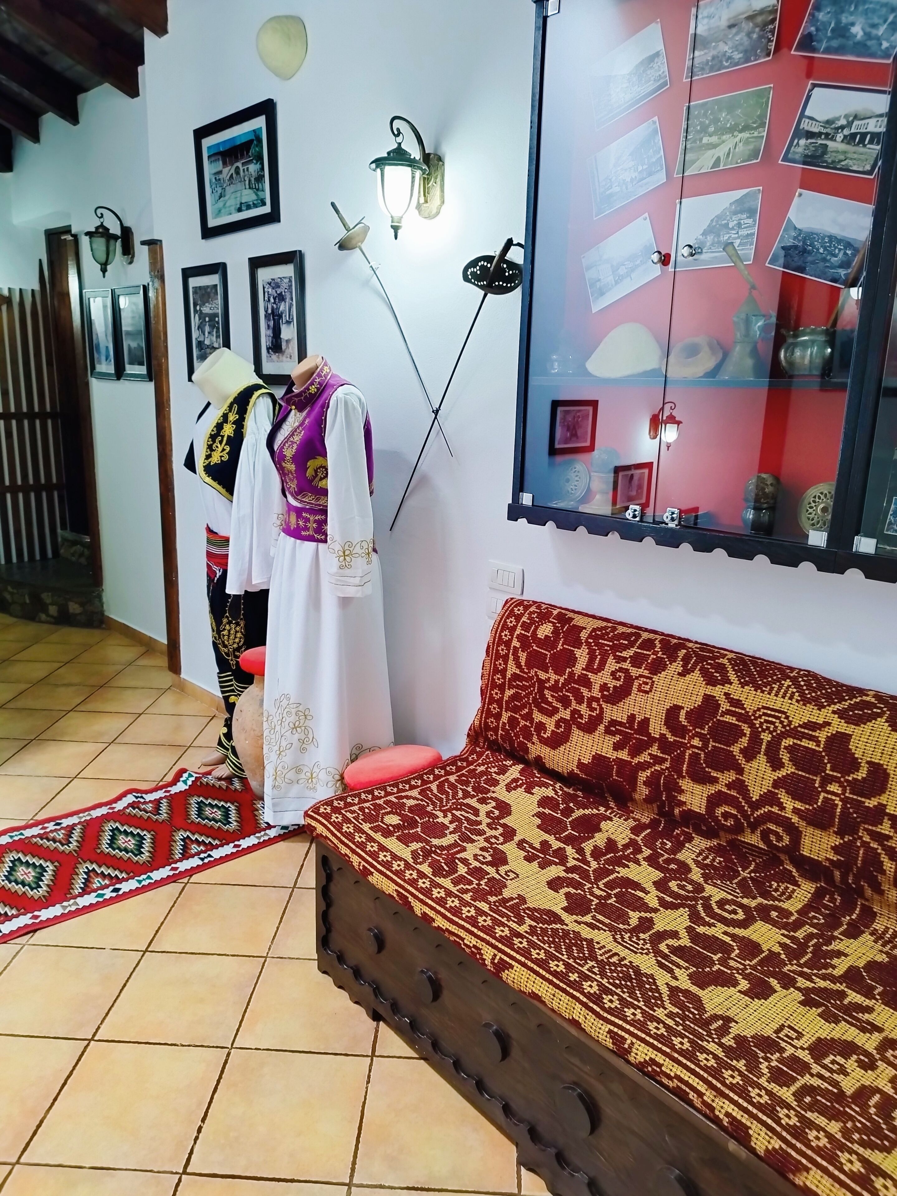 Una habitación con un sofá rojo estampado, un maniquí vestido con traje tradicional, y una pared decorada con cuadros enmarcados y una vitrina de cristal.