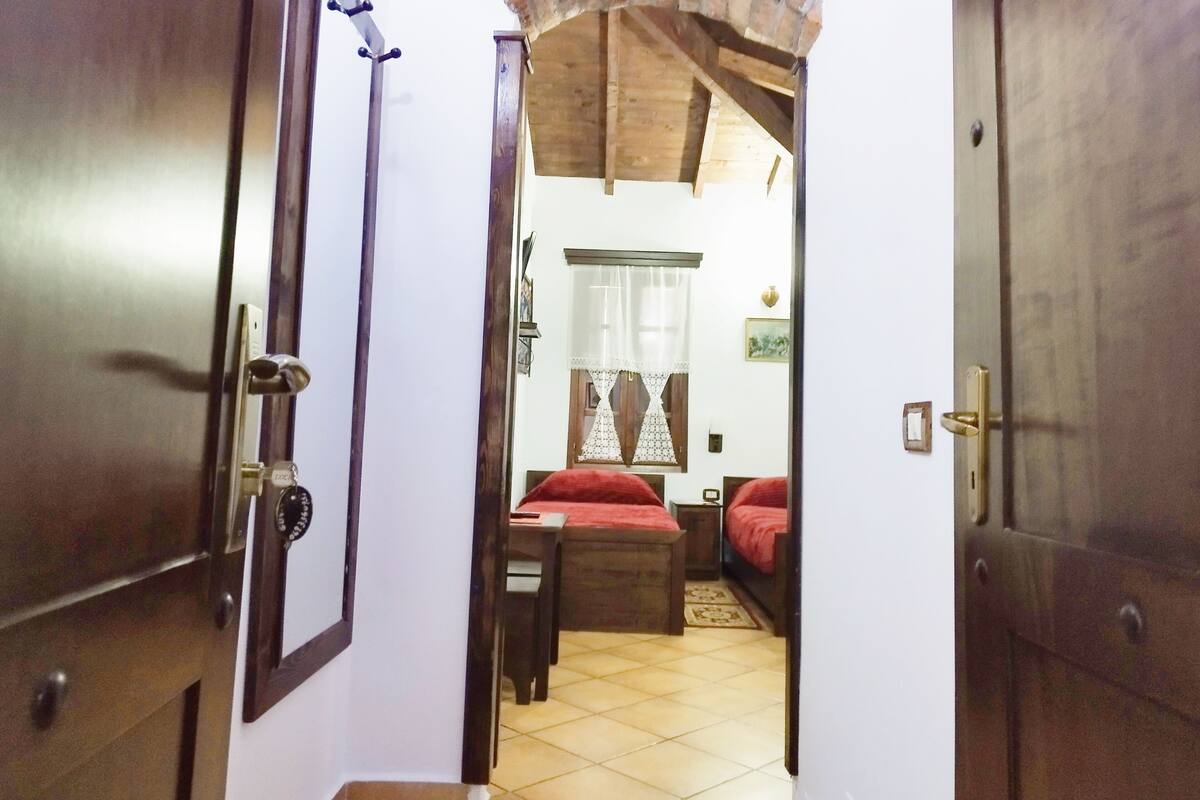 A room with a wooden ceiling, a bed with red bedding, a small table, and a chair.