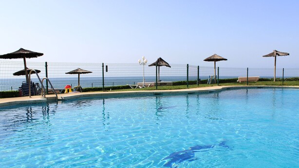Uma piscina com água azul cristalina, rodeada por uma cerca e vários guarda-sóis de palha.