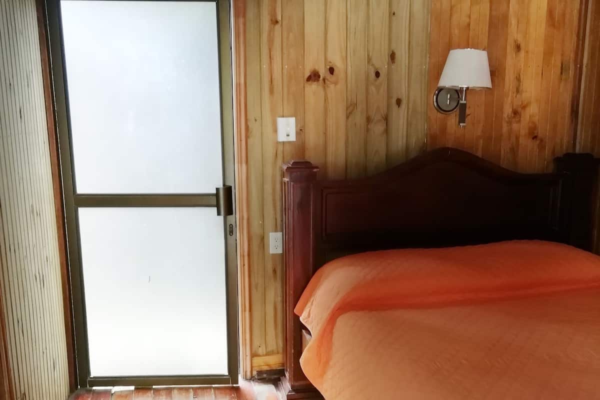 Quarto de madeira com cama, ar condicionado, luminária na parede e porta que dá para o exterior.