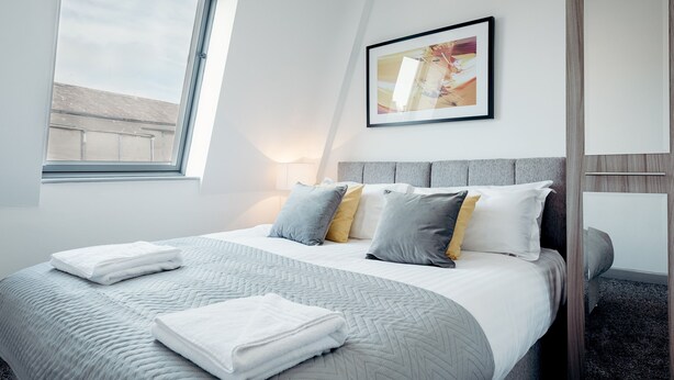Una habitación de hotel moderna con una cama grande, una ventana, un cuadro abstracto enmarcado y un espejo.