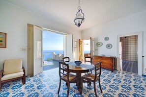 Un coin repas avec une table et des chaises, un luminaire suspendu et une vue sur la mer par une porte ouverte.