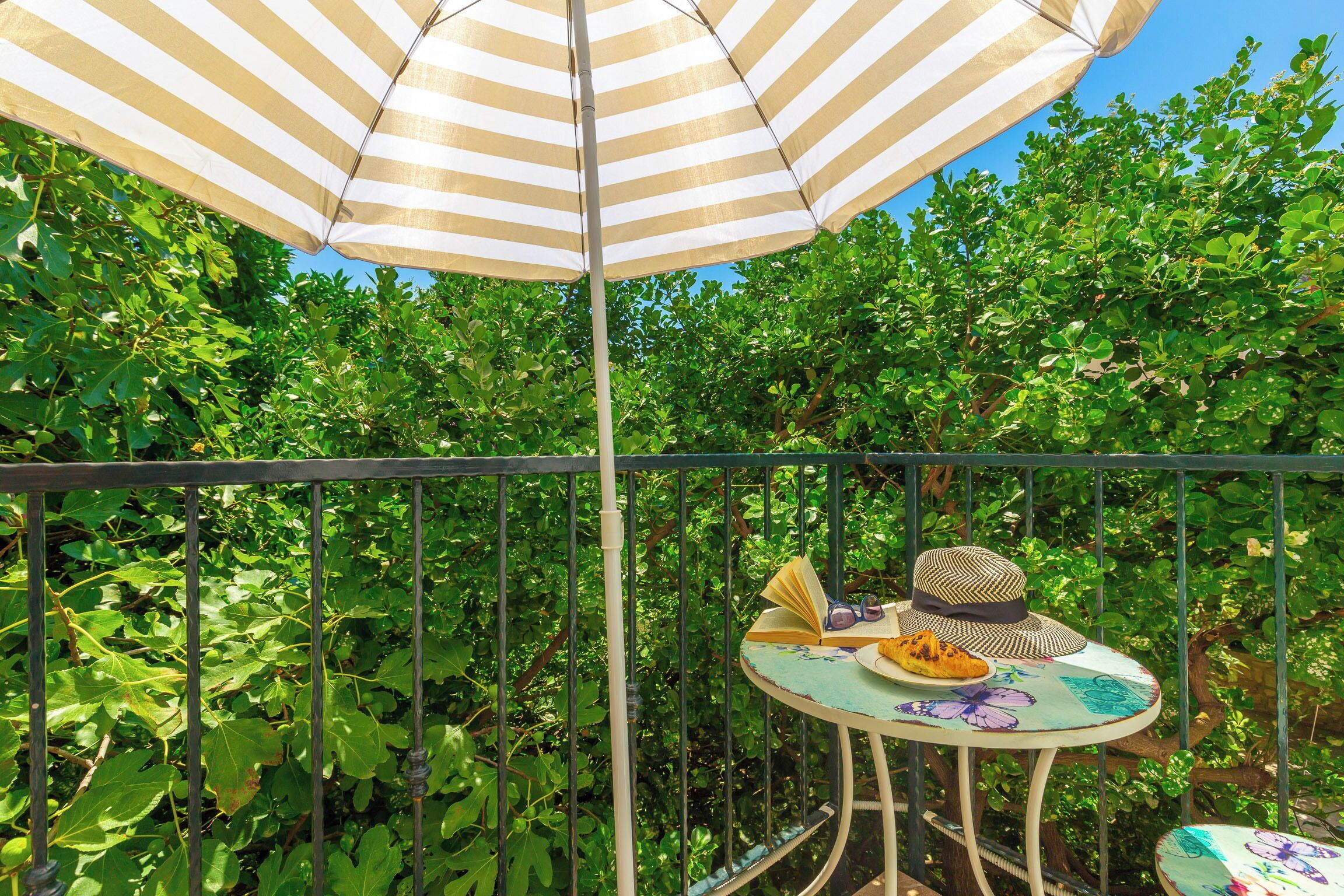 Een balkon met een gestreepte parasol, een tafel waarop een hoed, zonnebril en een boek liggen, omgeven door weelderig groen.