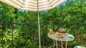 Uma varanda com um guarda-sol listrado, uma mesa com chapéu, óculos de sol e um livro, cercada por vegetação exuberante.
