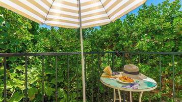 Een balkon met een gestreepte parasol, een tafel waarop een hoed, zonnebril en een boek liggen, omgeven door weelderig groen.