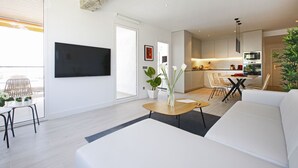 Een moderne woonkamer met een witte bank, een salontafel en een tv aan de muur bevestigd. Er is een eethoek met een tafel en stoelen, en een keuken op de achtergrond.