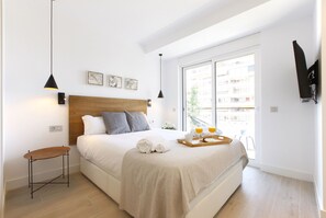 Une chambre moderne avec une tête de lit en bois, une table de chevet, un lit recouvert d’une couverture beige et un plateau avec des verres et un livre.