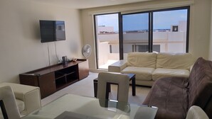 Ein modernes Wohnzimmer mit einem Glastisch, einem beigen Sofa, einem braunen Sofa, einem an der Wand montierten Fernseher und einer Schiebetür aus Glas, die auf einen Balkon führt.
