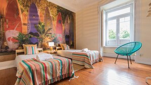 Ein Zimmer mit zwei Betten, einem Wandgemälde von Surfbrettern, einem Fenster mit Aussicht und einem türkisfarbenen Sessel.