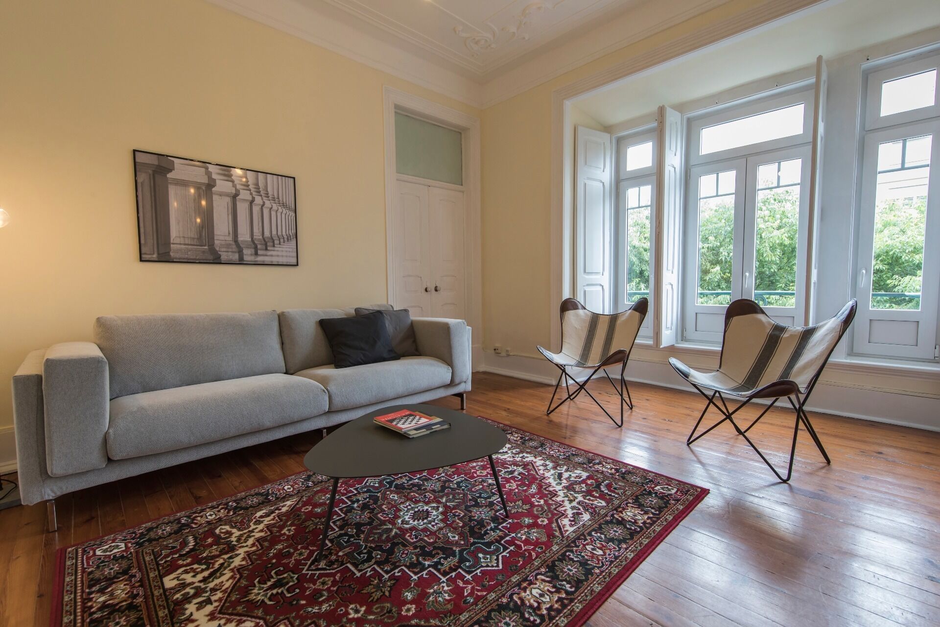 Ein Wohnzimmer mit grauem Sofa, einem gemusterten Teppich, einem Couchtisch und zwei Sesseln. Es gibt ein großes Fenster mit vielen Scheiben und eine Tür mit Glasfenster.