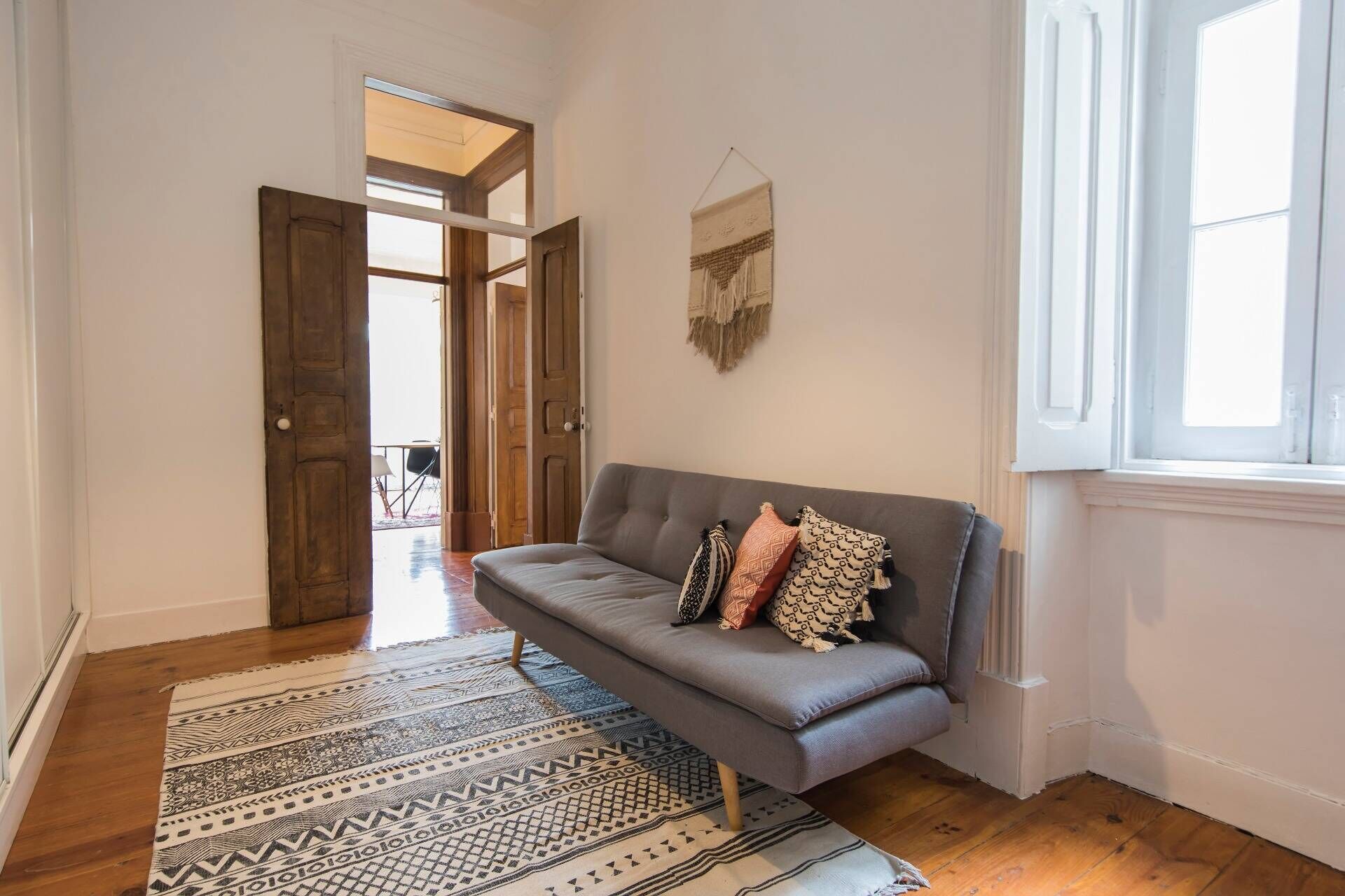 Una habitación con un sofá gris, alfombra estampada y una ventana con molduras blancas.