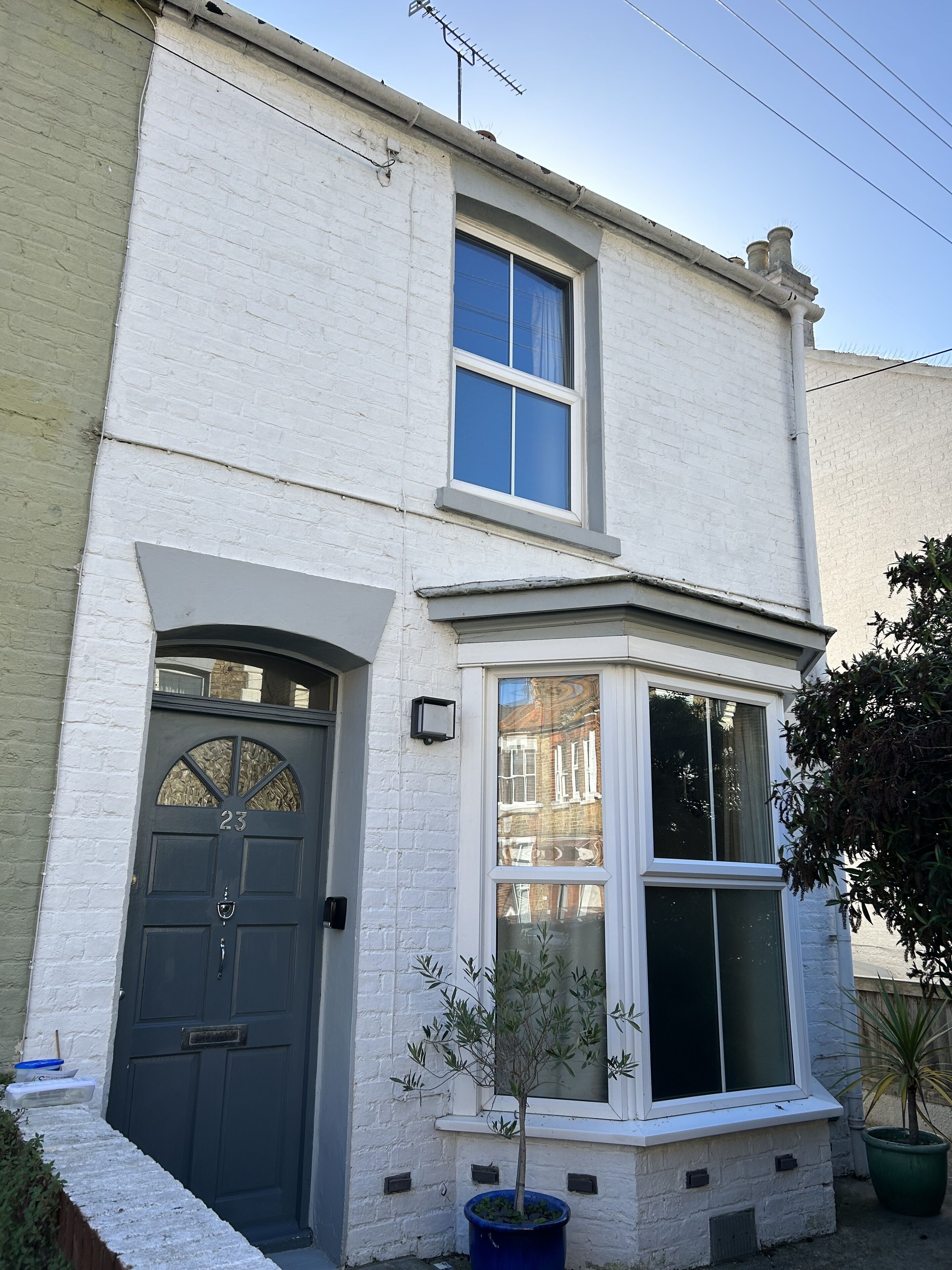 A two-story white brick house with a dark grey front door, a bay window, and a small tree in a blue pot.