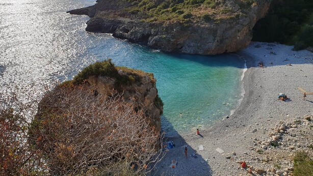 Un paysage côtier avec une plage rocheuse, des eaux turquoise et limpides, et un petit bâtiment situé sur une colline.