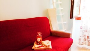 A red sofa with a tray of snacks and a bottle, a wooden floor, and a white chair in a bright room with a window. - My House of Milan 19 (Milano)