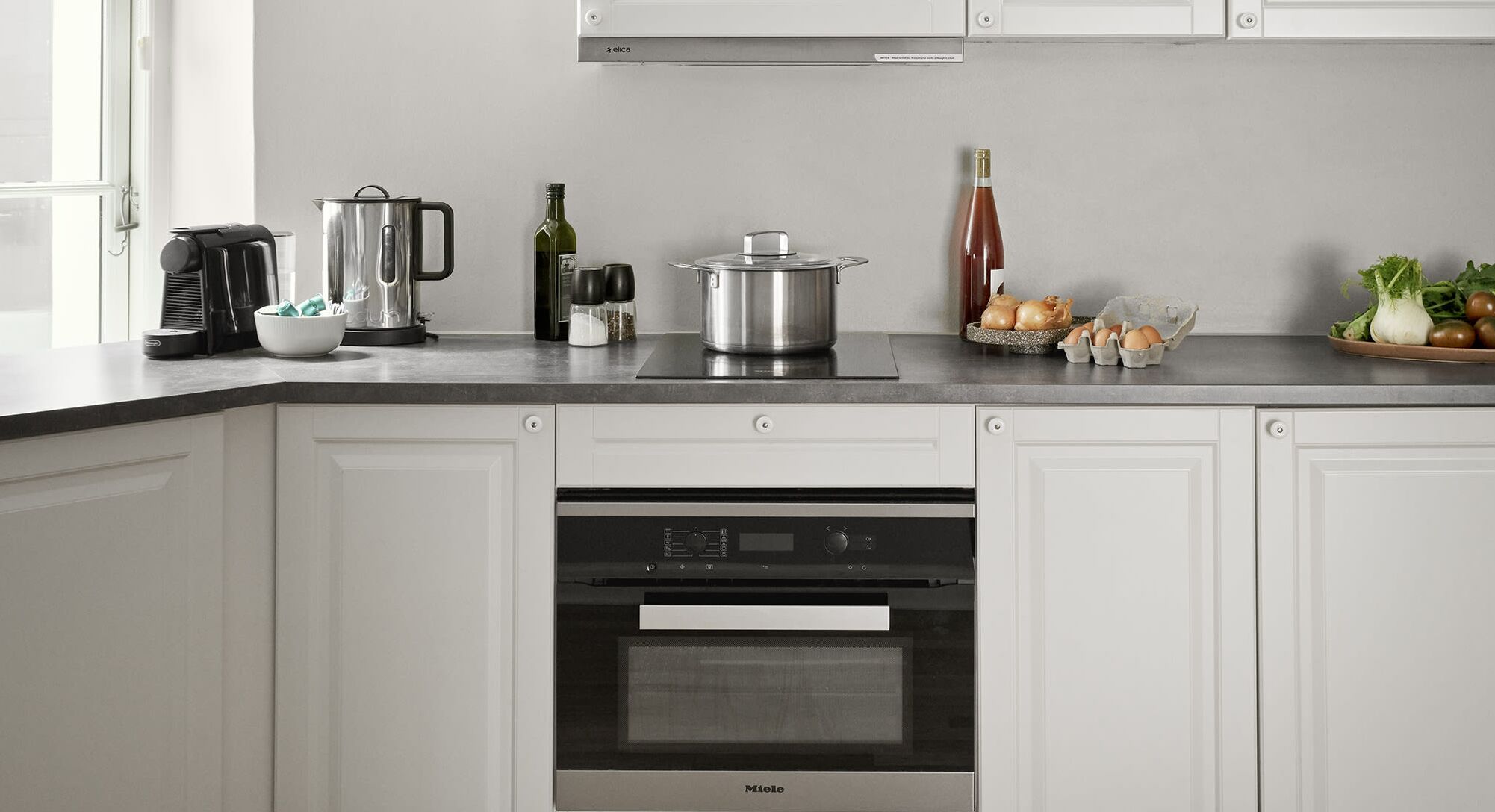 Una cocina moderna con horno empotrado, cafetera, hervidor, una botella de vino y productos frescos en la encimera.