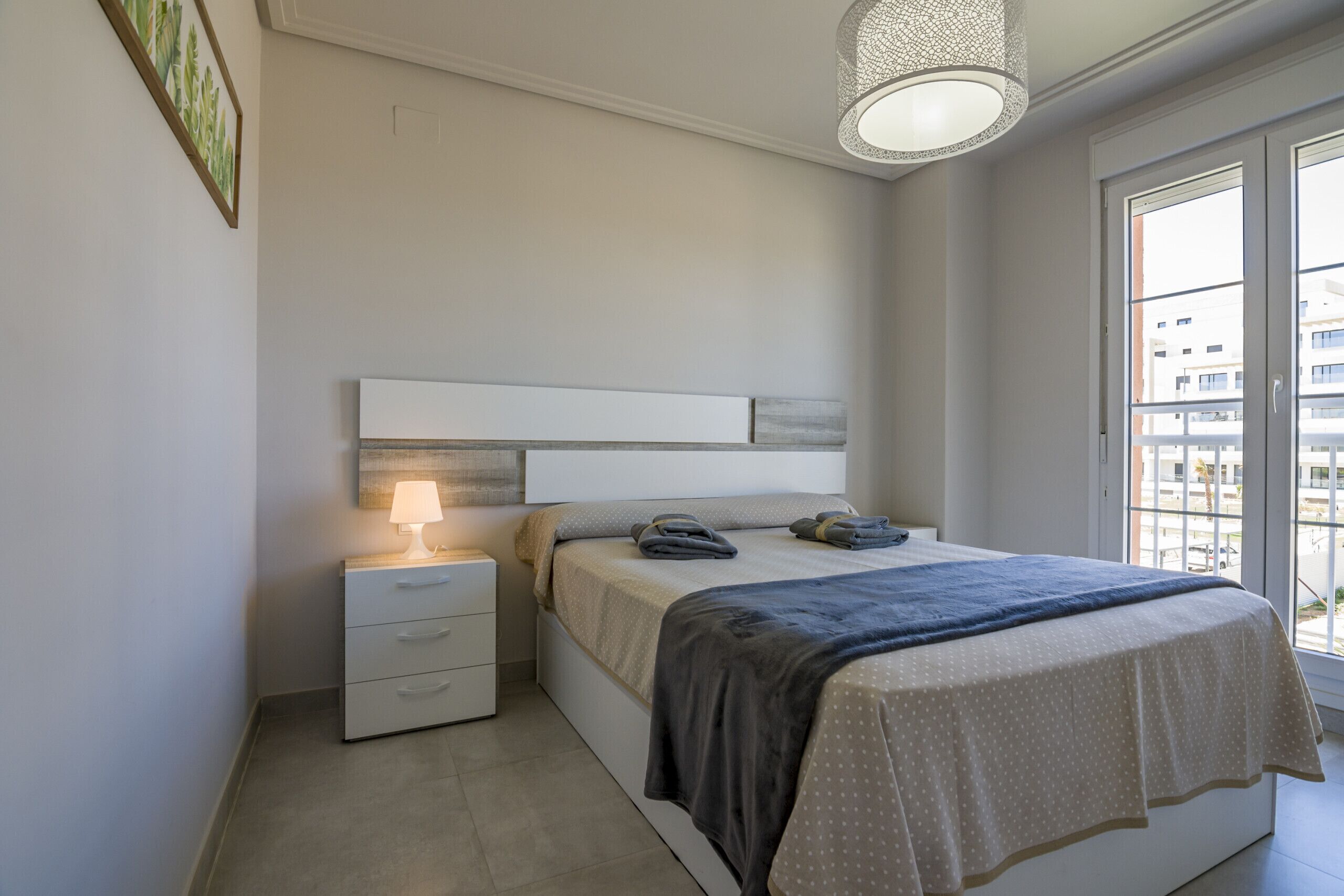Habitació amb llit, taulell de nit, làmpada i vista a edificis per la finestra.