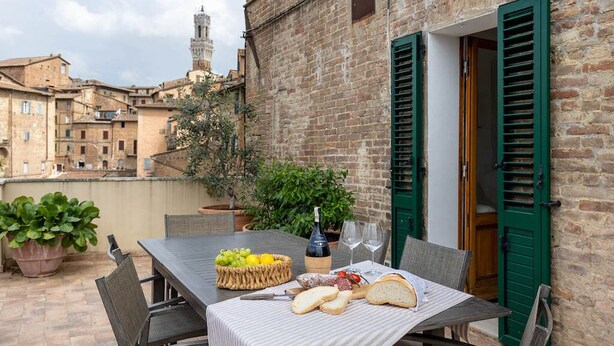 Une table dressée à l’extérieur, avec une bouteille, des verres, du pain et un panier de fruits.