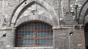 Une porte en bois à deux battants, surmontée d’une fenêtre à grilles métalliques, est encastrée dans un bâtiment en briques où l’on peut voir des conduits électriques et une petite grille d’aération.