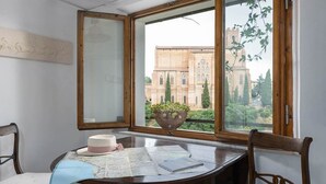 Une pièce dotée d’une grande fenêtre donnant sur un bâtiment historique, d’une table en bois sur laquelle sont posées des cartes, et d’un vase rempli de fleurs.