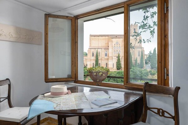 A room with a large window overlooking a historic building, a wooden table with maps, and a vase with flowers. - HOUSE WOW panoramic terrace *.* (Siena)