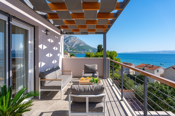 A balcony with a sofa, a table with a plant, and a view of the sea and mountains.