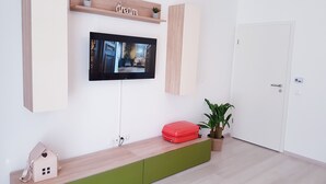 Ein modernes Wohnzimmer mit einem an der Wand montierten Fernseher, Holzregalen, einem kleinen hausförmigen Gegenstand und einem roten Koffer.