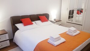 Ein ordentlich bezogenes Bett mit weißer und orangefarbener Bettwäsche, zwei rote Kissen und ein Stapel gefalteter weißer Handtücher auf einem orangefarbenen Bettüberzug.